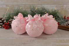 three pink balls with bows and pearls on them sitting in front of pineconis