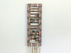 a jewelry organizer hanging on the wall next to a white wall with pink flowers and gold chains