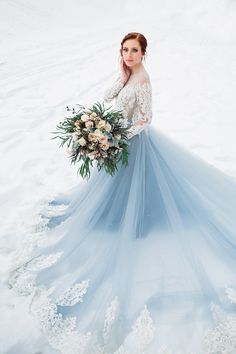 a woman is standing in the snow wearing a blue wedding dress and holding a bouquet