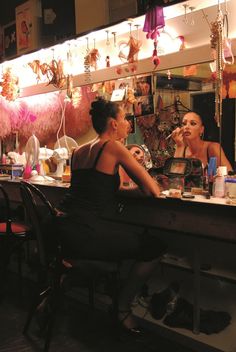 two women sitting at a table in front of a mirror with lights hanging from the ceiling