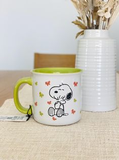 a coffee mug with a snoopy cartoon on it sitting next to a white vase