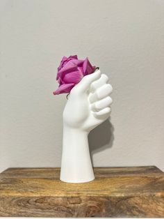 a white hand holding a pink rose on top of a wooden table next to a wall