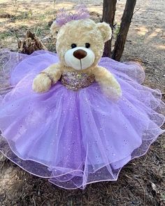 a teddy bear wearing a purple dress sitting on the ground in front of a tree