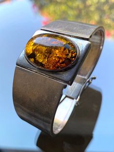 This is a vintage sterling silver bracelet with a large oval amber setting. It has an amazing patina on it and the amber glows in the sun with a rich deep honey hue. The sides of the bracelet are sturdily hinged to the silver setting plate, and the clasp is reminiscent of old jewelry. Watch the video to see the clasp in action. We think it is very old judging by the clasp and the patina and style. The inside tab of the clasp has a "YS" hallmark and "925". Measurements:  -Length- Inside length measurement 6 1/2"L  -Width- 1 3/8"W at the widest point, which is the silver rectangular setting for the amber. It is 1/2"W at the clasp.  -Weight- 1.307oz, or 37.05g Note: As this is pre-loved and not quite brand new, it has some minor fine scratches, but this is common with vintage sterling silver Vintage Brown Jewelry With Polished Finish, Vintage Amber Jewelry With Polished Finish, Vintage Amber Bangle Jewelry, Vintage Baltic Amber Jewelry For Formal Occasions, Vintage Amber Bangle Bracelet, Vintage Modernist Jewelry, Jewelry Watch, Length Measurement, Modernist Jewelry