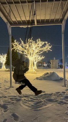 a person is swinging on a swing in the snow at night with christmas lights behind them