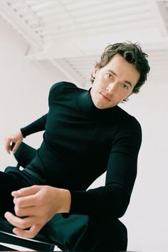 a young man sitting on top of a table in front of a white wall and wearing black pants