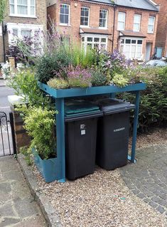 two black trash cans sitting next to each other on the side of a road with plants growing out of them