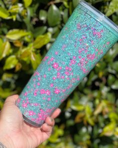 a hand holding a blue and pink glittered tumbler in front of some bushes