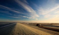 a truck driving down a dirt road next to the ocean under a blue sky with wispy clouds