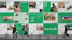 two people walking past a wall covered in green and white advertisement boards that read wework