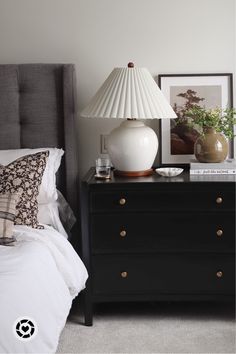 a white lamp sitting on top of a black dresser next to a bed with pillows