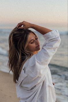 a woman standing on top of a beach next to the ocean holding her hair up