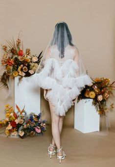 the back of a woman wearing a white wedding dress and veil with flowers behind her