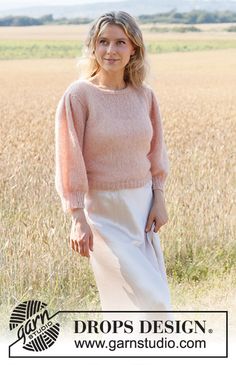 a woman standing in the middle of a field wearing a pink sweater and white skirt