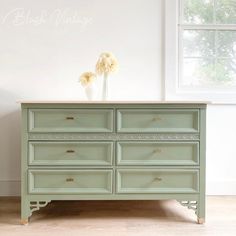 a green dresser sitting in front of a window with white flowers on top of it