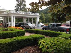 cars parked in front of a white gazebo surrounded by greenery and flowers on a cloudy day