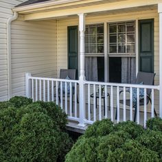 two chairs on the front porch of a white house with green shutters and bushes