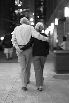 an older couple walking down the sidewalk in front of a building at night, back to back