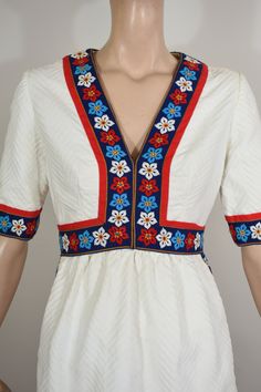 "This is such a great Oscar de la Renta boutique dress. The dress is vintage 60's. It is a white cotton matelasse fabric. The dress is trimmed with a embroidered blue ribbon that has red, turquoise, and white flowers with gold/ yellow centers. There is red ribbon trim running next to flower trim. This decoration runs around the neck, sleeve cuffs, waist hem and creates an apron shape on the front skirt. The cut is a V neck, fitted waist and bust, 3/4\" sleeve, and an Aline skirt with hip side po White Bohemian Vintage Dress For Garden Party, White Fitted Peasant Dress, White V-neck Vintage Dress, Vintage White V-neck Dress, White Retro V-neck Dress, White Vintage V-neck Dress, White Retro Vintage Dress With Floral Print, 1970s Style White Vintage Dress For Spring, Retro White V-neck Vintage Dress