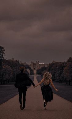 two people holding hands walking down a path in the middle of a park on a cloudy day