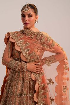 Pakistani Bridal Dress in Peach Lehenga Pishwas Style is an embellished attire that will give you a head-turning look on the most important day of your life. Lavish designs, embroidery, and ornaments give a chic look to this beautiful Pishwas Dress. Pakistani Bridal Pishwas: Pakistani Bridal Pishwas in an alluring peach shade has premium net fabric. The Pishwas Frock is gracefully emblazoned with zardosi, dabka, floral designs, and naqshi. The Traditional Pishwas is embellished with intricate designs, silver and gold details, giving it a flawless finishing look. Bridal Lehenga: The Bridal Lehenga has premium quality tissue fabric and it looks perfect with the heavily embellished Pishwas Frock. The Lehenga in peach gold color has a huge flare and its borders are emblazoned with tilla, naqsh