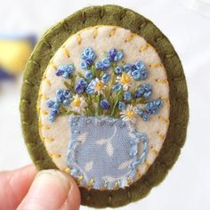 a hand holding up a small patch with blue flowers in it and a green frame