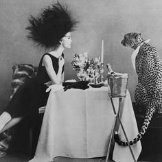 a woman sitting at a table with a cat on top of her head next to a vase