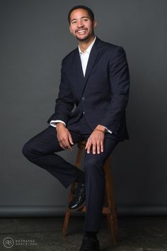 a man in a suit and tie sitting on a wooden stool smiling at the camera