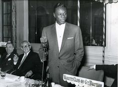 a black and white photo of a man in a suit speaking into a microphone while others look on