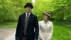 a man and woman dressed in period clothing walking down a dirt road with trees behind them