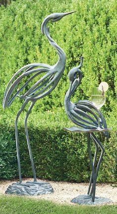 two metal birds standing next to each other on top of a lush green field with bushes