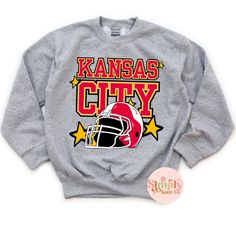 Youth Kansas City Sweatshirt, Kansas City Football Sweatshirt for Kids, Kansas City Shirt, Kansas City Gift, KC Football Crewneck Sweatshirt HOW TO ORDER: Select size from drop down menu Select shirt color from drop down menu Choose quantity MATERIALS:  Printed in shop with DTF (Direct to Film) and then pressed onto shirt. If unsure or in between sizing, I always recommend sizing up!!   Youth sweatshirt Gildan or Jerzee brand depending on stock. Unisex sizing, please refer to size chart  SHIPPING: I will do my best to process your order in a timely manner. I process them in the order I get them. Current processing time is 5-7 days. I use USPS shipping with tracking.  I will not be held responsible for any lost, damaged, or delayed packages once they are shipped out. If an item shows delive Winter Fan Apparel T-shirt With Logo Print, Winter Team Spirit T-shirt With Graphic Print, Winter Team Spirit Graphic T-shirt, Long Sleeve T-shirt With Name Print For Sports Events, Winter Fan Gear Graphic T-shirt, Football Season Cotton Graphic Sweatshirt, Cotton Graphic Print Sweatshirt For Football Season, Football Crewneck, City Sweatshirt