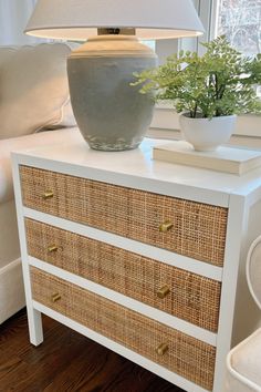 a white dresser with wicker drawers and a lamp on top in front of a window