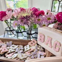 pink and purple flowers are in vases on a table next to cookies that spell out the word bake