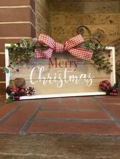 a merry christmas sign with red and white plaid bows on it sitting on a brick floor