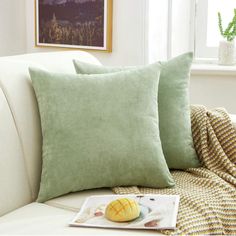 two green pillows sitting on top of a white couch next to a book and potted plant