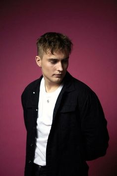 a young man in black jacket and white t - shirt standing against a pink background