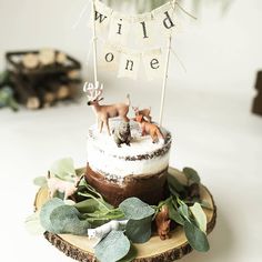 a cake decorated with deer figurines and greenery on a wooden slice that says wild one