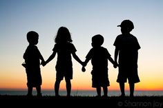 three children holding hands while standing in front of the ocean at sunset, silhouetted against an orange and blue sky