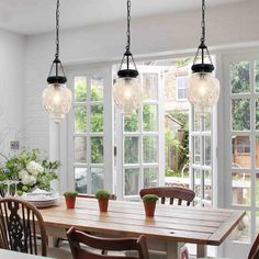 a dining room table with chairs and lights hanging from it's ceiling, in front of large windows