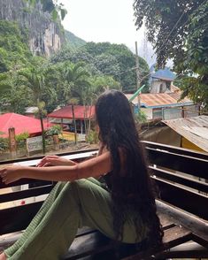 a woman sitting on top of a wooden bench next to a lush green forest covered hillside