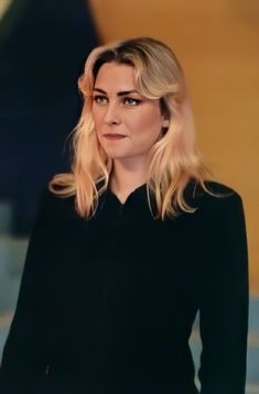 a woman with blonde hair wearing a black dress and looking off to the side while standing in front of a yellow wall