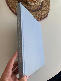 a person holding up a blue book on top of a white table next to a straw hat