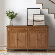 a wooden sideboard with two framed pictures on top and a plant in vase next to it