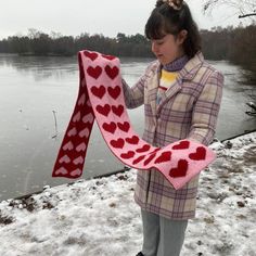 a woman holding up a scarf with hearts on it in the snow near a lake