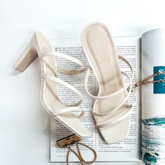 A pair of ivory strappy heels that have a square toe. These shoes are pictured on a white background with gold jewelry and a magazine. Spring Sandals With 4-inch Heel And Square Toe, Summer Block Heels With 4-inch Square Toe, Trendy Cream Sandals With Padded Heel, Cream Ankle Strap Block Heels For Summer, Spring 4-inch Square Toe Heels, Cream Open Toe Heels For Summer, Summer Cream Open Toe Heels, Chic Strappy Heels With Stacked Heel, Trendy Beige Sandals With 4-inch Heel