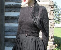 a woman wearing a black dress and hat standing in front of a log cabin with her hands on her hips