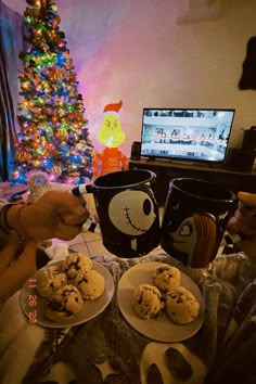 two people sitting at a table with plates of cookies and coffee mugs in front of a christmas tree