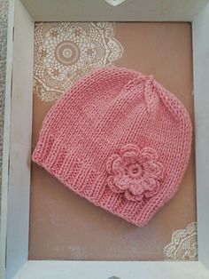 a pink knitted hat with a flower on it in a white box next to doily