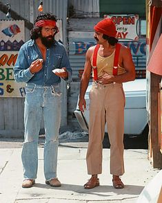 two men are standing on the sidewalk eating food and looking at each other while one man is wearing a bandana